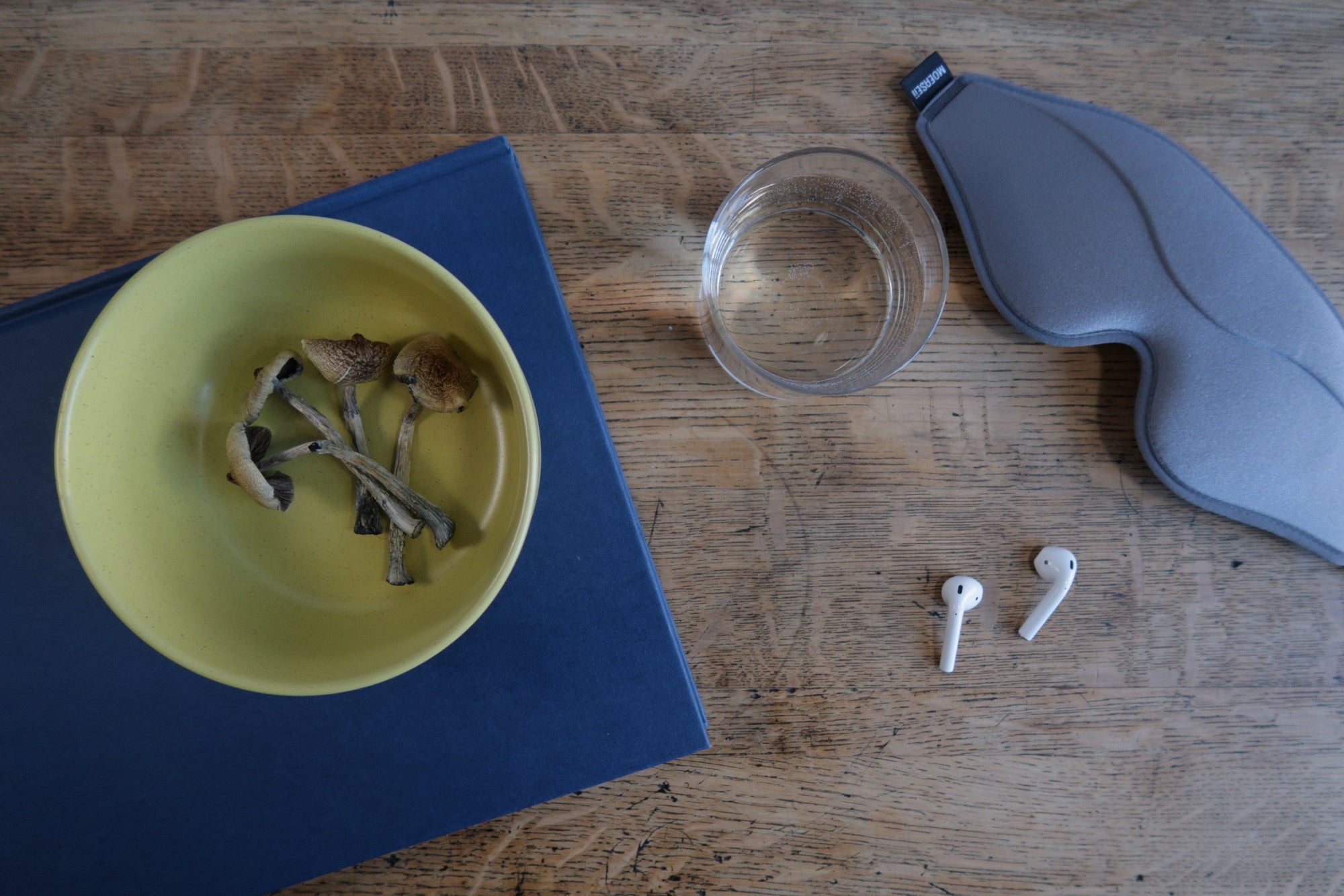 Yellow bowl with mushrooms, clear glass, white earbuds, and gray sleep mask on a wooden surface